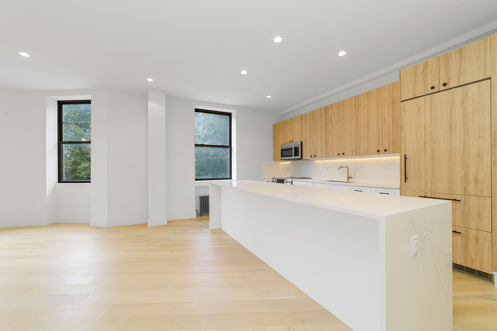 Studio, One, Two, & Three-Bedroom Apartments in Upper West Side Manhattan, NY - 125 Riverside - Living Room and Kitchen with Light Wood-Style Flooring, 2 Windows with Black Trim, White Walls, a Marble Kitchen Island, Light Wood-Style Cabinets, Stainless Steel Appliances, and Marble Backsplash